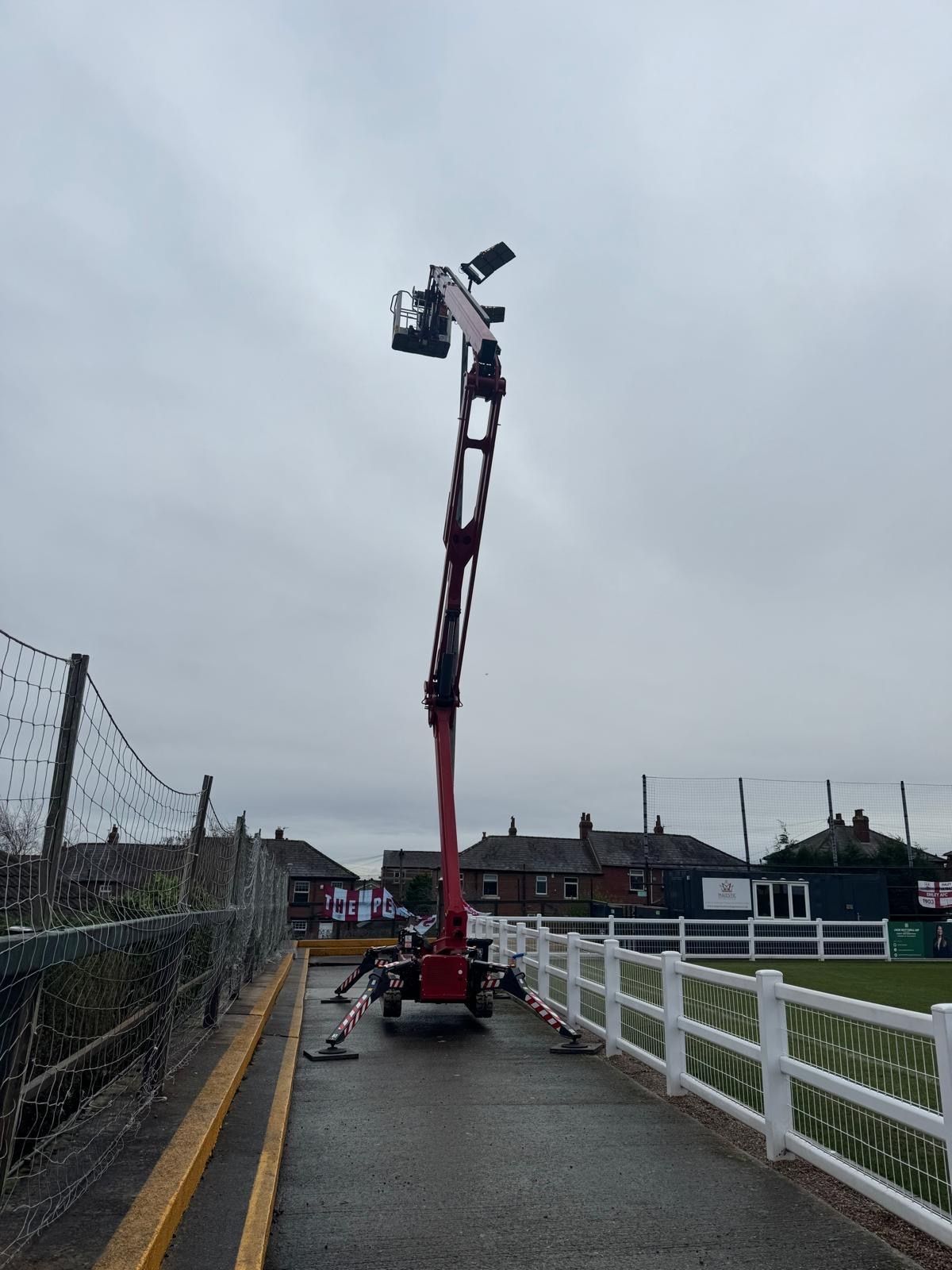 Red spider lift set on a narrow path beside a sports pitch with its boom raised